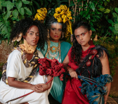Três integrantes do Clarianas com roupas coloridas, flores e cordas enfeitando seus trajes.