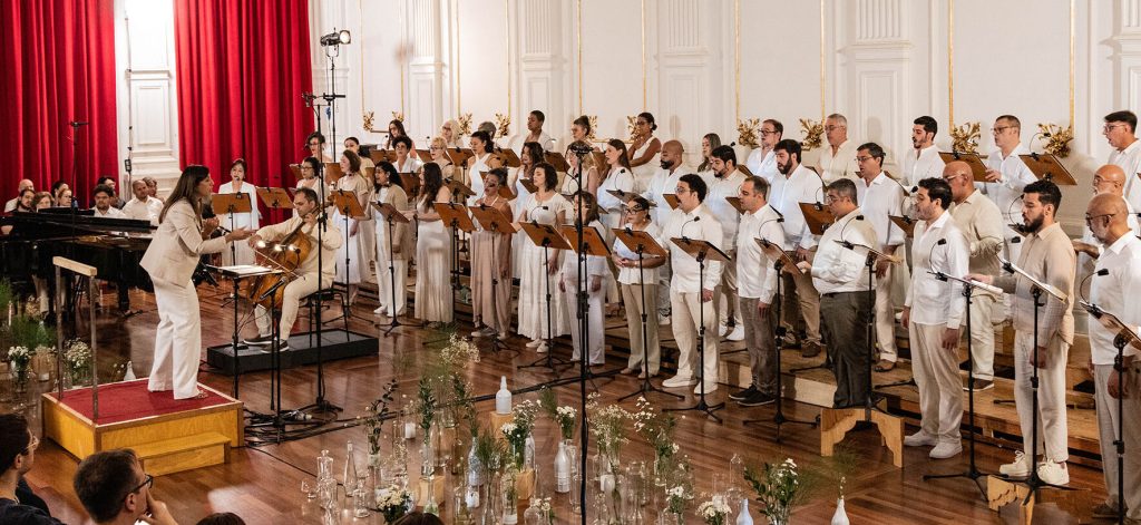 Na imagem o coral está posicionado com a maestra a frente, todos vestidos de branco.